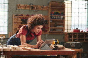 Young person with curly hair working on a tablet in a woodworking shop.
