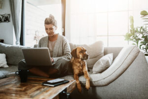 Woman working on laptop, sitting on couch next to a dog.